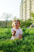 süßes kleines Mädchen, das an einem sonnigen Frühlingstag einen Kuss schickt, im Gras sitzt