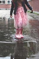 kleines Mädchen in Gummistiefeln, das nach dem Regen in einer kleinen Pfütze spielt