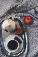 Tasse Kaffee, Mokkakanne auf silbernem Tablett und Äpfel auf der grauen Strickdecke, Draufsicht, winterlicher gemütlicher Hintergrund