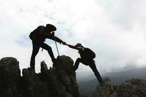 Silhouette von zwei männlich Wanderer Klettern oben Berg Cliff und einer von Sie geben Portion Hand. Menschen Portion Und, Mannschaft Arbeit Konzept. foto