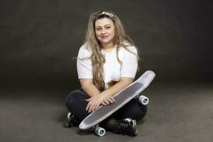 schön Fett Frau mit ein Skateboard auf ein grau Hintergrund sieht aus beim das Kamera und lächelt. foto