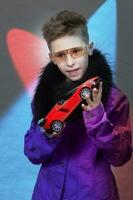 ein modisch Junge ist halten ein Spielzeug Auto im seine Hände, auf ein mehrfarbig Studio Hintergrund. ein schön Kind im futuristisch Brille träumend von ein teuer Wagen. Träume von ein Kind. ein Reich Kind. foto