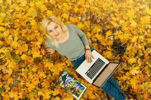 süß Frau mit Laptop im das Herbst Park. Schönheit Natur Szene mit bunt Laub Hintergrund, Gelb Bäume und Blätter beim fallen Jahreszeit. Herbst draussen Lebensstil. glücklich lächelnd Frau auf fallen Blätter foto