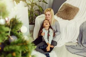 jung Mutter und ihr wenig Tochter magisch Weihnachten Geschenk durch ein Weihnachten Baum im gemütlich Leben Zimmer im Winter foto