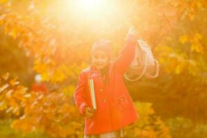 wenig süß Mädchen läuft entlang Herbst Park und Tritte oben gefallen Blätter. Schülerin mit ein Rucksack geht von Schule.die Konzept von Herbst, Schule, lernen, Ausbildung, Kindheit, zurück zu Schule. foto