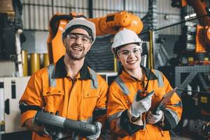 glücklich jung Ingenieur Arbeiten zusammen Bedienung Mannschaft mit Roboter Schweißen im Automatisierung Fabrik. Mann und Frau lächelnd Arbeiter im Sicherheit passen Mitarbeiter Arbeiter im automatisiert Technologie Herstellung Industrie. foto