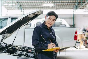 glücklich asiatisch Mechaniker jung männlich Mitarbeiter Arbeiter Arbeiten mit Auto Motor Vorderseite Kapuze Auto Bedienung prüfen Tuning Motor im Garage foto