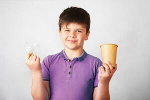 lächelnd Junge hält ein Plastik und ein Papier Trinken Tasse im das Hände. Beförderung von Nichtbenutzung von Plastik. Null Abfall Konzept foto