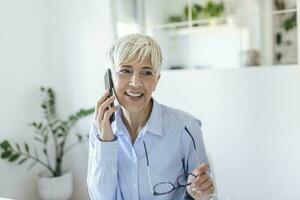 Senior Frau mit ihr Telefon. glücklich Frau entspannend beim heim, sie ist reden auf ihr Smartphone foto