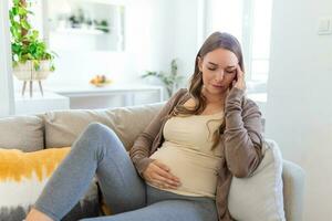 schwanger jung Erwachsene Frau ruhen auf Sofa beim heim, Gefühl nicht wohl. jung schwanger Frau hat litt von Kopfschmerzen Sitzung auf das Sofa. Schwangerschaft Symptome foto