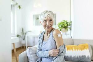 Senior Frau suchen glücklich nach bekommen Impfung. reifen Frau mit Pflaster auf ihr Arm nach Empfang covid-19 Impfung Sitzung beim Zuhause foto