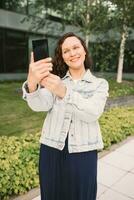 jung Frau im Weiß Denim Jacke nehmen Selfie auf Straße. Büro Gebäude im das Hintergrund foto