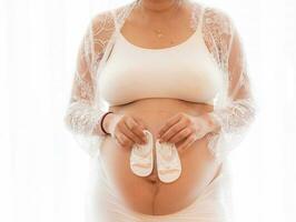 Nahansicht schwanger Bauch mit süß winzig Baby Schuhe. Konzept von Schwangerschaft, Gynäkologie, Vorbereitung. schön schwanger Frau im Hemd im Weiß Hintergrund. Frau halten groß schwanger Magen. foto