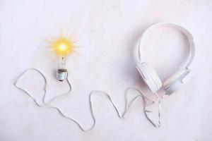 Glühbirne mit Kopfhörern auf weißem Tischhintergrund, Ideeninnovations- und Inspirationskonzept, neue Technologie in der Zukunft