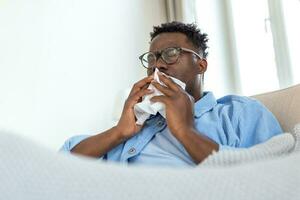 krank afrikanisch jung Mann bedeckt mit Decke weht Laufen Nase habe Fieber gefangen kalt Niesen im Gewebe sitzen auf Sofa, krank allergisch schwarz Mann haben Allergie Symptome Husten beim heim, Corona 19 foto