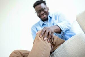 Foto von afrikanisch Mann Sitzung auf ein Sofa im das Leben Zimmer beim Zuhause und berühren seine Knie durch das Schmerzen während das Tag. Mann massieren seine schmerzlich Knie.