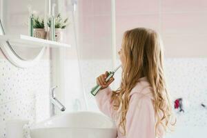 süß Kind Mädchen im ein Rosa Frottee Dressing Kleid Bürsten ihr Zähne im Vorderseite von das Badezimmer Spiegel im das Morgen foto