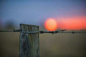 ein Zaun Post mit mit Widerhaken Draht und ein Sonnenuntergang im das Hintergrund foto