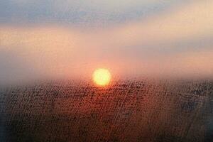 defokussiert verschwommen Himmel Hintergrund. Sonnenuntergang durch nass Fenster Glas, Kondensation Textur. Orange Sonne hell Platte im Center, das dunkel Horizont unten ihnen. abstrakt Hintergrund mit Platz zum Ihre Text. foto