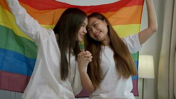 Porträts von glücklich asiatisch Lesben Paar Sitzung mit Regenbogen Flagge Sitzung Bett neben das Fenster. heiter zwei jung Erwachsene Frauen Leben zusammen mit Beziehung Konzept. lgbtq Lebensstil Stolz Monat foto