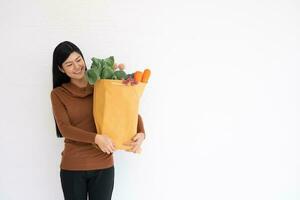 glücklich asiatisch Frau ist lächelnd und trägt ein Einkaufen Papier Tasche nach das Kurier von das Lebensmittelgeschäft kam zu liefern seine Waren beim heim. Konzept von Supermarkt Lieferung zum ein Neu Lebensstil foto