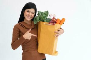 glücklich asiatisch Frau lächelnd und Punkt das Finger zu ein Papier Tasche und trägt ein Einkaufen Tasche nach das Kurier von Lebensmittelgeschäft kam zu liefern beim heim. Konzept von Supermarkt Lieferung zum ein Neu Lebensstil. foto