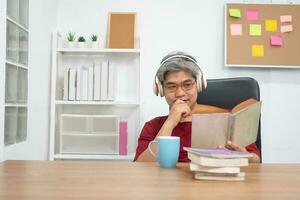 entspannen Alten asiatisch Mann lesen ein Buch und Hören zu Musik- im Zuhause Büro. foto