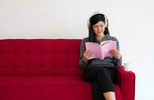 süß asiatisch Frau ist Sitzung auf das Sofa im das Morgen und lesen ein Buch während Hören zu Musik- im das Leben Zimmer foto