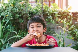 asiatischer süßer Junge im roten Hemd, der glücklich sitzt und Pizza isst. foto