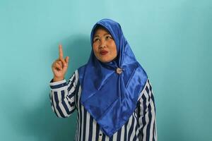 ein aufgeregt mittleren Alters asiatisch Frau im ein Blau Hijab und gestreift Hemd ist zeigen ihr Finger oben beim das Kopieren Raum, isoliert Über ein Blau Hintergrund foto