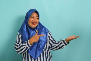 mittleren Alters asiatisch Frau im Blau Hijab und gestreift Hemd zeigen zu ihr öffnen Palme. Stehen gegen ein Blau Hintergrund. foto