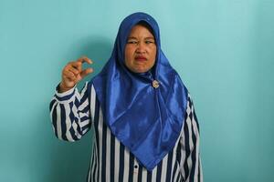 ein unzufrieden mittleren Alters asiatisch Frau im ein Blau Hijab und gestreift Hemd zeigt an ein klein Geste, suchen enttäuscht beim etwas sehr winzig im Größe, während Stehen gegen ein Blau Hintergrund. foto