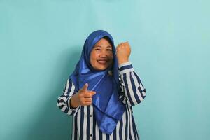 ein aufgeregt mittleren Alters asiatisch Frau im ein Blau Hijab und gestreift Hemd zeigt an ein Daumen hoch und ein okay Zeichen, symbolisieren Erfolg und ausdrücken Genehmigung, während Stehen gegen ein Blau Hintergrund. foto