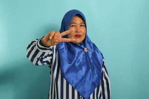 mittleren Alters asiatisch Frau im Blau Hijab und gestreift Hemd Anzeigen ein Frieden oder Sieg Geste mit ihr Hand. Stehen gegen ein Blau Hintergrund. foto