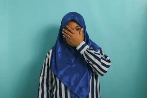 überrascht mittleren Alters asiatisch Frau im Blau Hijab und gestreift Hemd Abdeckung Mund, starren beim Kamera im Erstaunen. isoliert auf Blau Hintergrund. foto