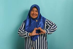 ein lächelnd mittleren Alters asiatisch Frau, im ein Blau Hijab und gestreift Shirt, macht ein Herz Geste mit ihr Hände, ausstrahlen Glück und Pflege, isoliert auf Blau Hintergrund foto