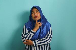 ein nachdenklich Mitte alt asiatisch Frau, im ein Blau Hijab und gestreift Shirt, denkt und Sorgen Über ein Frage, erscheint besorgt und nervös mit Hand auf kinn, Stehen gegen Blau Hintergrund foto