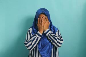 ein erschrocken aber neugierig mittleren Alters asiatisch Frau im Hijab versteckt sich ihr Gesicht, schließt ihr Augen mit ihr Hände, und späht durch ihr Finger während Aufpassen ein Grusel Film. isoliert auf ein Blau Hintergrund. foto
