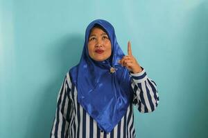 ein aufgeregt mittleren Alters asiatisch Frau im ein Blau Hijab und gestreift Hemd ist zeigen ihr Finger oben beim das Kopieren Raum, isoliert Über ein Blau Hintergrund foto