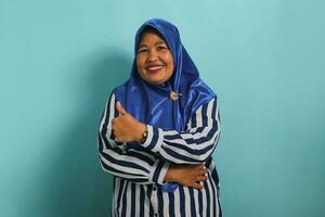 ein aufgeregt mittleren Alters asiatisch Frau im ein Blau Hijab und gestreift Hemd zeigt an ein Daumen hoch und ein okay Zeichen, symbolisieren Erfolg und ausdrücken Genehmigung, während Stehen gegen ein Blau Hintergrund. foto