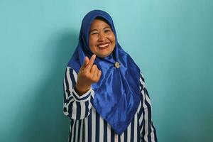 ein mittleren Alters asiatisch Frau, im ein Blau Hijab und gestreift Shirt, macht ein Mini Herz Hand Zeichen oder Koreanisch Finger Herz Zeichen, isoliert auf ein Blau Hintergrund. foto