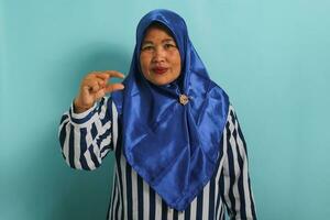 ein unzufrieden mittleren Alters asiatisch Frau im ein Blau Hijab und gestreift Hemd zeigt an ein klein Geste, suchen enttäuscht beim etwas sehr winzig im Größe, während Stehen gegen ein Blau Hintergrund. foto