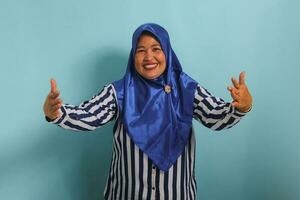 ein freundlich mittleren Alters asiatisch Frau, im ein Blau Hijab und gestreift Shirt, Spreads aus ihr Hände mit ein lächeln, eifrig warten Umarmungen und herzlich einladend Du, isoliert auf ein Blau Hintergrund foto