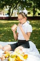 ein süß Mädchen Schülerin Getränke Saft mit ein Stroh von ein umweltfreundlich Tasse beim ein Picknick auf ein Decke im das Park. Schule Mahlzeiten. Vertikale Aussicht foto