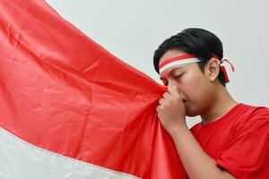 Porträt von respektvoll asiatisch Mann im T-Shirt mit Stirnband Band auf Kopf, küssen rot und Weiß Flagge von Indonesien. isoliert Bild auf grau Hintergrund foto