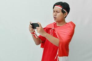 Porträt von attraktiv asiatisch Mann im T-Shirt mit rot Weiß Band auf Kopf mit Flagge auf seine Schulter wie ein Mantel, spielen Spiel auf Handy, Mobiltelefon Telefon mit wütend Ausdruck. isoliert Bild auf grau Hintergrund foto