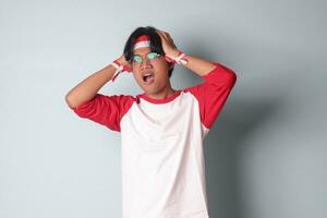 Porträt von attraktiv asiatisch Mann im T-Shirt mit rot und Weiß Band auf Kopf, haben ein Migräne, berühren seine Tempel. Kopfschmerzen Konzept. isoliert Bild auf grau Hintergrund foto