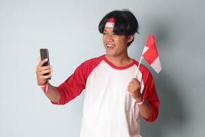 Porträt von attraktiv asiatisch Mann im T-Shirt mit rot und Weiß Band auf Kopf, nehmen ein Bild von selbst während halten Indonesien Flagge. isoliert Bild auf grau Hintergrund foto
