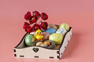 Ostereier in einem natürlichen Nest auf einem Holztablett, Blumen und ein dekoratives Huhn auf rosa Hintergrund