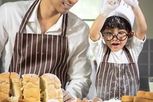 asiatischer Junge trägt eine Brille, neckt Papa, der mit Weißmehl kocht, der Brotteig knetet, lehrt Kinder das Backen von Zutaten Brot, Ei auf Geschirr im Küchenlebensstil glückliches Lernen mit der Familie foto